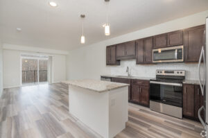 A modern kitchen and living area with light wood flooring. Features dark wood cabinets, granite countertops, and stainless steel appliances. Two pendant lights hang above the island. A sliding glass door leads to a balcony with a view.