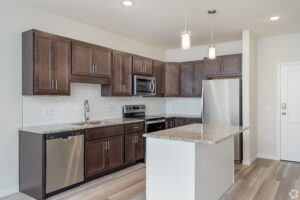 Modern kitchen with dark wood cabinets, stainless steel appliances, and a granite countertop island. White subway tile backsplash and light wood flooring add contrast. Two pendant lights hang above the island.