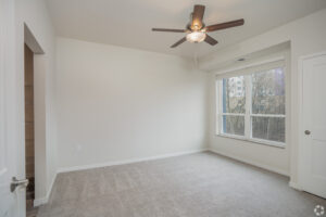 An empty room with carpeted flooring, a ceiling fan, and a large window letting in natural light. The walls are painted white, and the room features a partially open door on the right. Another open door leads to a small closet on the left.