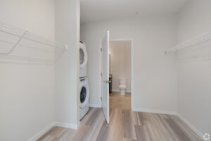 A laundry room with a stacked washer and dryer on the left. The room has light wood flooring and white walls. Wire shelving units are mounted on both sides. An open door leads to a bathroom with visible toilet.