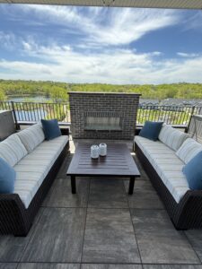 Two patio sofas with striped cushions and blue pillows face each other across a dark wood table on a tiled balcony. A brick fireplace stands behind the table. The balcony overlooks a scenic view of trees and a river under a partly cloudy sky.