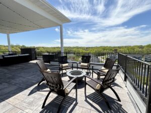 Rooftop patio with wooden chairs arranged around a fire pit on a tiled floor. Overhead, string lights are attached to a partial roof. The view includes trees and a neighborhood under a blue sky with scattered clouds.