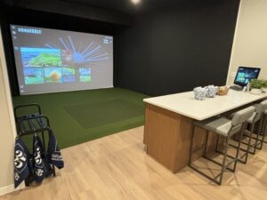 Indoor golf simulation room with a large screen displaying a virtual golf course. A golf bag is to the left, and a bar area with high chairs and baskets of golf balls is on the right. The floor is wooden with green turf near the screen.