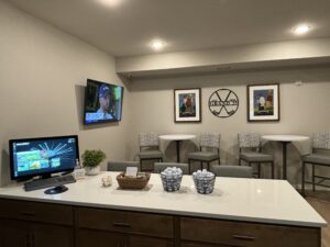 A recreation room with a computer on a desk to the left, a mounted TV showing a person, and framed pictures on the wall. There are high tables and chairs against the wall, with baskets of golf balls on the counter in the foreground.