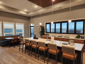 Modern kitchen interior with a large island counter featuring six cushioned stools. Pendant lights hang above. To the left, a breakfast nook with grey cushions. Large windows provide natural light, overlooking a scenic view.