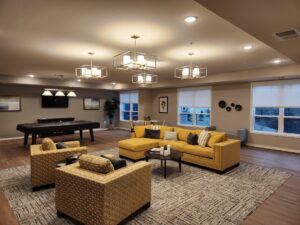A modern living room with yellow sofas and two armchairs on a patterned rug. The room features a pool table, a mounted TV, and large windows with roller blinds. Ceiling lights with geometric fixtures illuminate the space.