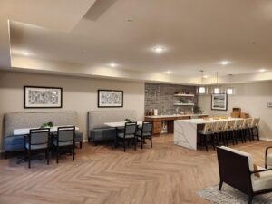 A modern dining area with wooden floors, featuring several booths with grey seating, tables, and chairs. A bar with stools sits on the right, under pendant lights. Artwork adorns the walls, and small potted plants decorate the tables.