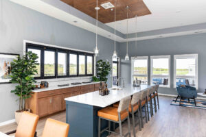 Modern kitchen and dining area with a long island featuring tan leather barstools. There are wood cabinets, a row of windows, pendant lights, and potted plants. Large windows in the background overlook a scenic view.