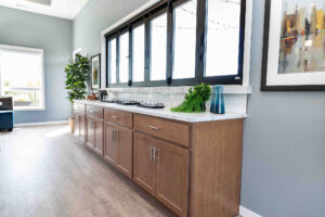 Modern kitchen interior with light wood cabinets and a white countertop. A row of large windows above cabinets provides natural light. Potted plant and decorative items adorn the counter, with framed art on the wall. Hardwood floor adds warmth.