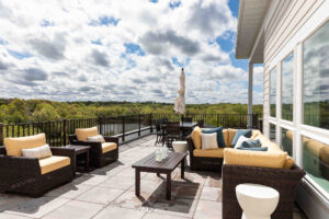 Rooftop patio with wicker furniture, cream cushions, and a central wooden table. An umbrella stands beside a dining set. The view shows a forest and a cloudy sky. Large windows are to the right.