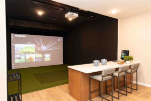 A home golf simulator setup featuring a large projection screen displaying a golf course. In front, there is a white counter with four stools, a small tablet, and baskets of golf balls. The room has light wood flooring and recessed lighting.