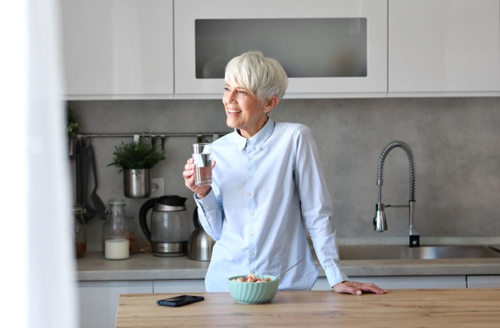 An older adult stands in their kitchen, smiling while drinking water and enjoying a bowl of fruit.